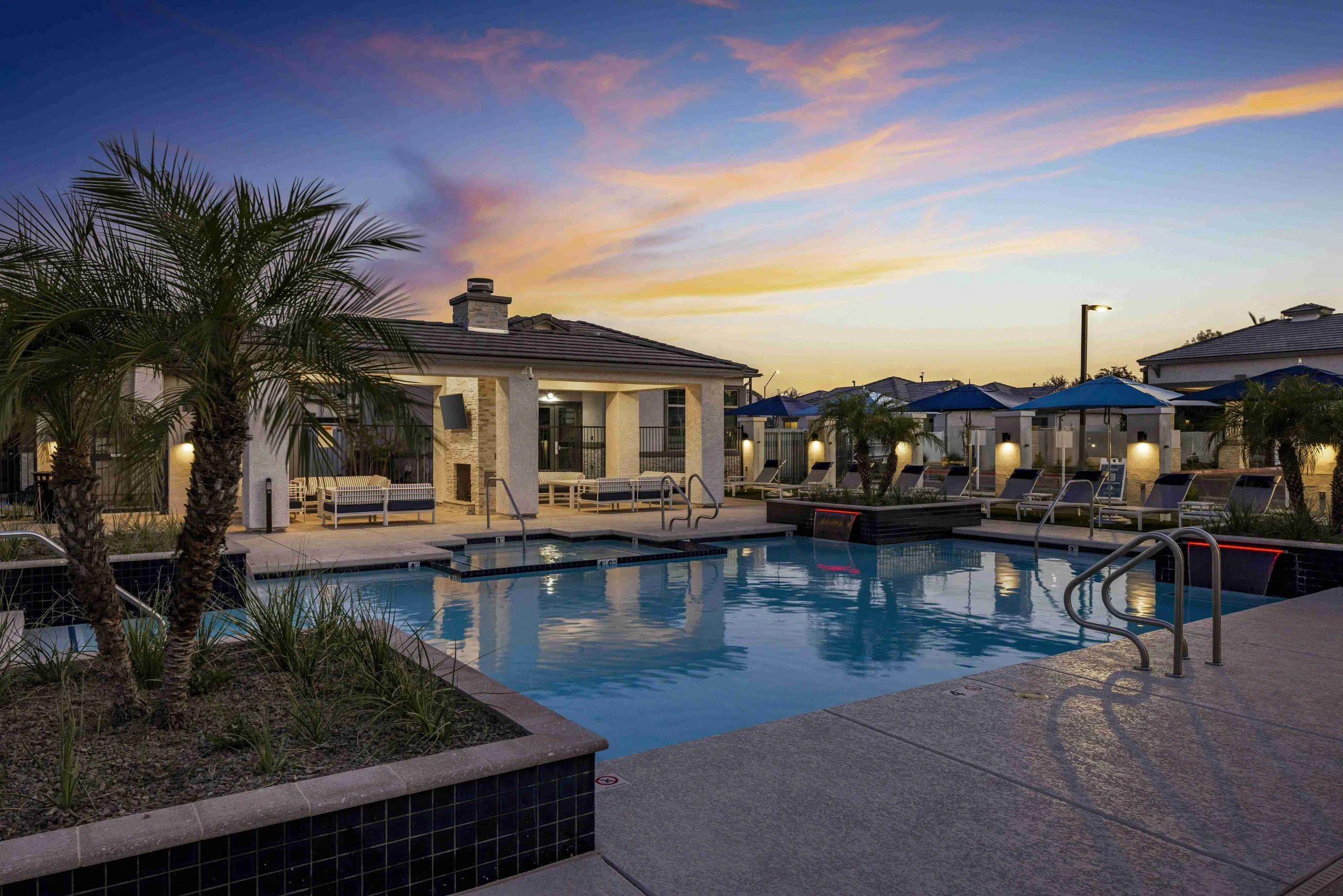 Pool and spa deck during sunset with water features, outdoor covered pergola, and lawn chairs on grass.