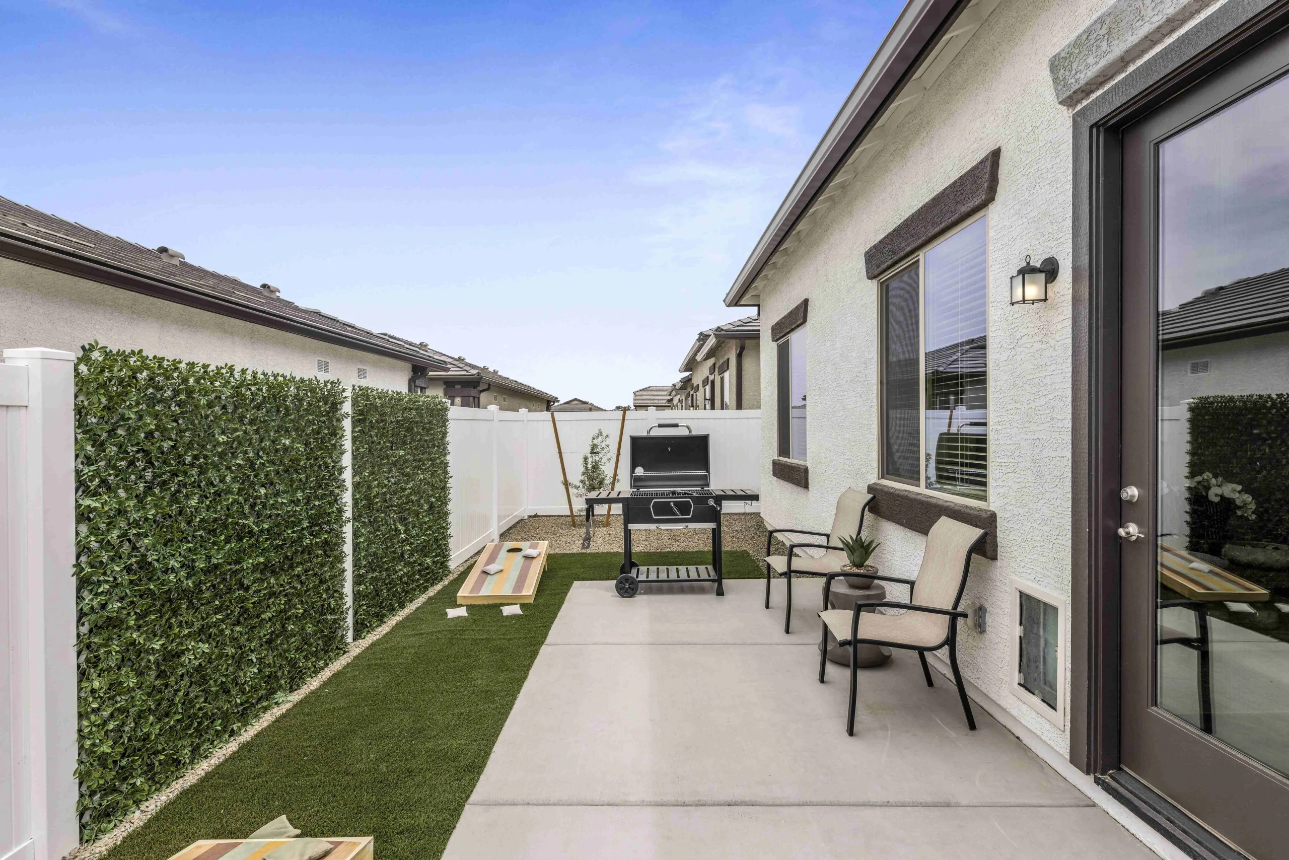 Backyard with seating, side table, BBQ, and cornhole with turf, gravel, a planted tree and cement patio.